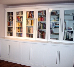 A large free-standing bookcase, incorporating glass doors to the top to display books and storage units to the bottom. Built in M.D.F. with a high gloss white painted finish. Made specifically to slot into the space available and built in separate sections for easy removal.
